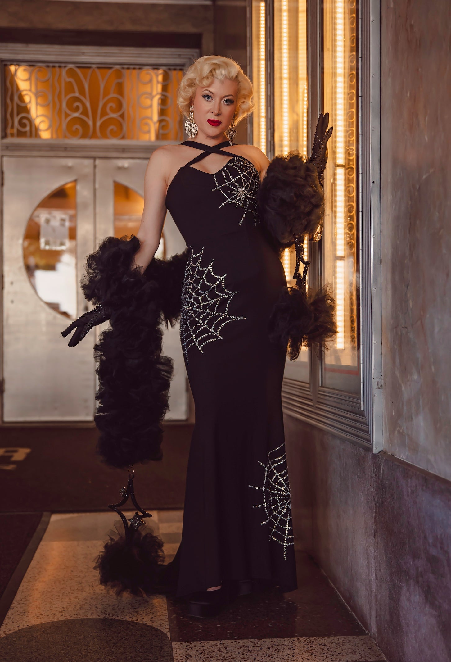A person with platinum blonde hair poses indoors in the Glamour Ghoul Gown by MURDER QUEEN X ELLE REBEL, a long black gothic dress with sparkling spiderweb designs, styled with black gloves and a feathered shawl near art deco doors and warm lights.